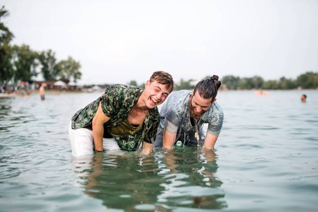 Wet young men friends at summer festival, standing in lake. Wet young men friends at summer festival, standing in lake and looking at camera. model released Copyright: xx Uprising_2019_1831