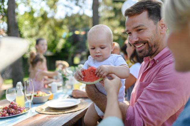 Cute baby eating watermelon at a summer garden party. Dad holding the baby boy in his arms and smiling. Cute baby eating watermelon at a summer garden party. Handsome father holding the baby boy in his arms and smiling. model released