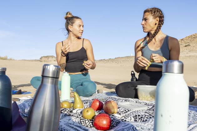 Two women enjoying a nutritious picnic on a beach mat, surrounded by fresh fruit and water bottles, engaged in a conversation with a clear blue sky above. Model Released JuanAizpuru_ID19654_582744_024 Copyright: xJuanxAizpurux