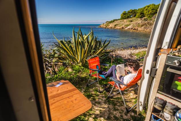 View from the sliding door of a motorhome standing on a pitch on the unpaved shore on the Mediterranean coast, a young woman sits on camping chairs and reads a book Sicily, Italy A motorhome stands on a pitch on the shore of the Mediterranean Sea