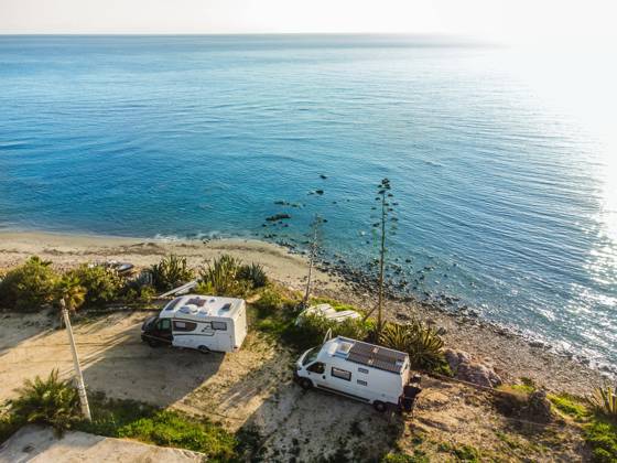 A camper van and an alcove motorhome are parked on a pitch on the unpaved shore on the Mediterranean coast, boats lie in front of them on the beach Sicily, Italy A motorhome is parked on a pitch on the shore of the Mediterranean Sea