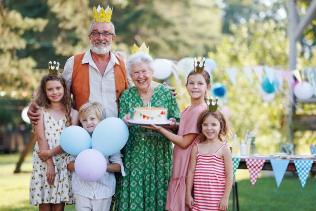 Garden birthday party for senior lady. Beautiful senior birthday woman holding cake and posing for pohoto with grandchildren and husband. Garden birthday party for senior lady. Beautiful senior birthday woman holding cake and posing for pohoto with grandchildren and husband. Birthday celebration with paper crowns. model released