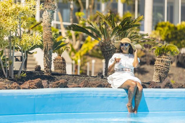 A smiling middle aged hispanic woman in a white dress and sunhat sits at the edge of a pool, holding a cocktail, surrounded by exotic plants. Model Released JuanAizpuru_ID20788_613331_028 Copyright: xJuanxAizpurux