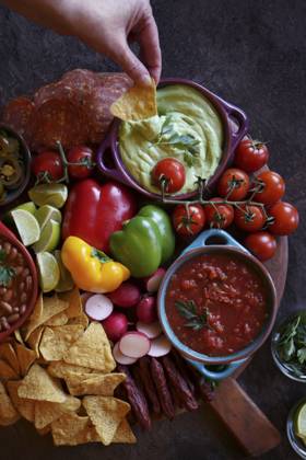 Mexican theme charcuterie board grazing platter with dips, salsa, beans, jalapenos, chorizo, and tortilla chips. Hand dipping guacamole. Mexican theme charcuterie board grazing platter with dips, salsa, beans, jalapenos, chorizo, and tortilla chips. Hand dipping guacamole. Copyright: xMilleflorexImagesx MIL190224-9