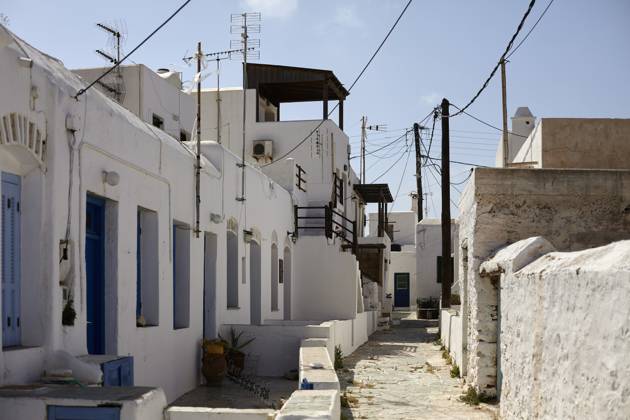 A narrow alley in a traditional Mediterranean village with white buildings and blue doors under a clear sky, with electrical wires crisscrossing overhead. Folegandros Greece bola01001 Copyright: xConnectxImagesx