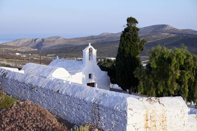 A serene landscape featuring a traditional white-washed church with a bell tower, surrounded by a stone wall, overlooking a hilly terrain with the sea in the distance at dawn. Folegandros Greece bola01005 Copyright: xConnectxImagesx