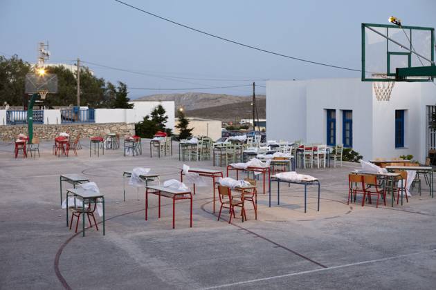 An outdoor setting at dusk featuring a basketball court with tables and chairs neatly arranged across it, under the glow of gentle lights, adjacent to a white building with blue trimmings. Folegandros Greece bola01016 Copyright: xConnectxImagesx