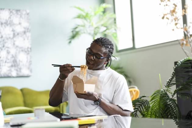A black man sits in a comfortable indoor setting, using chopsticks to eat noodles from a takeout box Model Released VictorCuenca_ID22085_646425_028 Copyright: xVictorxCuencax
