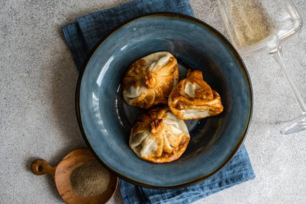 IMAGO Creative, Food, Dumplings A close-up view of traditional Georgian khinkali dumplings on a textured blue ceramic plate, accompanied by a wooden scoop of spices and a clear wine glass, set against a gray tabletop Model Released AnnaBogush_ID22388_654123_002 Copyright: xAnnaxBogushx