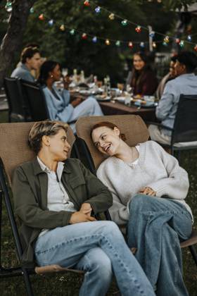 IMAGO Creative, Lifestyle, Feste im Garten #2 Female friends with legs crossed at knee sitting on chair and talking in back yard at social party Sweden, Stockholm, model released, property released Copyright: xMaskotx, MA112455