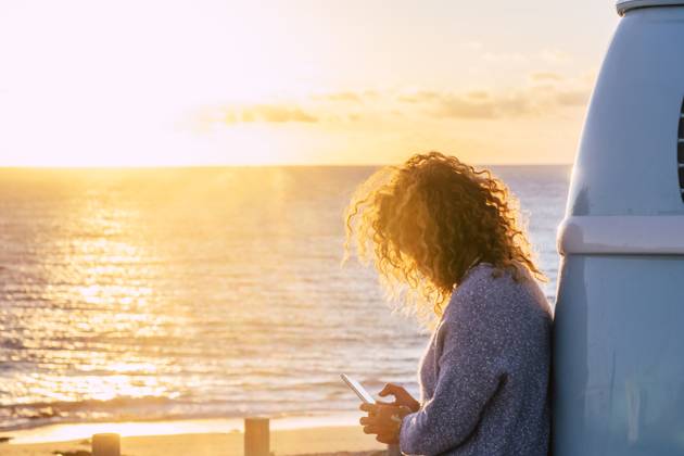 Woman using smart phone by beach at sunset in Fuerteventura, Canary Islands , 04.07.2019, model released, TTBB_ti0564_00080.jpg, Color Image,Horizontal,Focus On Foreground,Selective Focus,Outdoors,Sunset,Dusk,Fuerteventura,Canary Islands,Spain,Travel Destinations,Travel,One Person,People,40-44 Years,Mid Adult,One Mid Adult Woman Only,Only Women,Adult,Mid Adult Women,Caucasian Appearance,Personal Land Vehicle,Camper Van,Road Trip,Transportation,Getting Away From It All,Tourist,Beach,Sand,Coastline,Sea,Water,Blond Hair,Curly Hair,Sweater,Holding,Smart Phone,Communication,Sunbeam,Sunlight,Text Messaging,Mobile Phone,Wireless Technology,Cloud,Leaning Copyright: xFabioxCamandonax