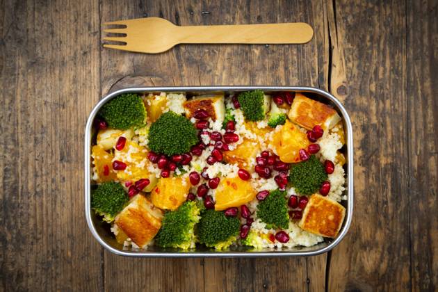 Stainless steel lunch box with couscous, broccoli, halloumi, oranges, chili dressing, and pomegranate seeds, on a wooden table with a wooden fork., LVF09377