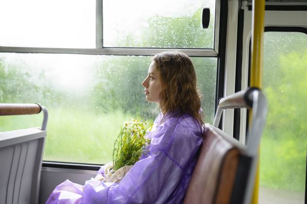IMAGO Creative, Jahreszeit Sommer, Regen, Sommerregen Creative Highlight Symbolbilder Woman in purple raincoat holding wild flowers sitting on a bus model released, Symbolfoto, VCF00804