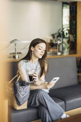 IMAGO Creative, Food, Trend, Iced Coffee A woman sits comfortably on a sofa in a cozy cafe, holding an iced coffee in one hand and a smartphone in the other, epitomizing relaxation and modern lifestyle. Model Released HannaYakimenko_ID30226_1019070_009 Copyright: xHannaxYakimenkox RECORD DATE NOT STATED