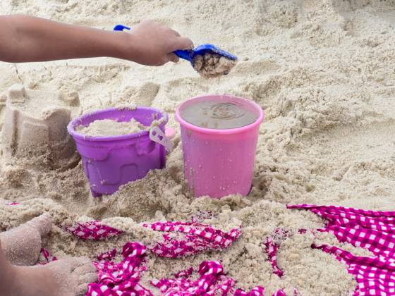 Child Building Sandcastles with Colorful Buckets at the Beach layful scene of a child s hand scooping wet sand into a bright pink and purple bucket using a blue plastic shovel. The sandy beach is scattered with footprints, sandcastles, and a red and white checkered fabric, enhancing the summer vacation vibe. This image captures childhood joy, outdoor fun, and beachside adventures, making it perfect for travel and lifestyle content., 95429814.jpg RECORD DATE NOT STATED