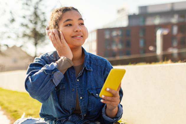 Girl Sitting on Ground Looking at Cell Phone A young girl with headphones is seated on the ground, engrossed in her cell phone screen. Her attention is solely focused on the device as she scrolls and types on the screen., model released, , 59453930.jpg RECORD DATE NOT STATED