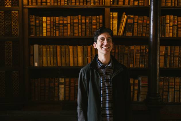 Young adult man smiling in front of book shelf in library Oxford, England, United Kingdom CR_WVNP251008B-1799229-01 ,model released, Symbolfoto