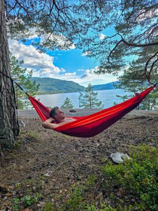 Entspannen in einer Hängematte an den ruhigen Gewässern Norwegens unter einem strahlend blauen Himmel A men enjoys a peaceful moment in a vibrant hammock, nestled between trees by Norways tranquil lake. The stunning landscape features lush greenery and distant mountains against bright skies. (License=RF) ,model released, Symbolfoto Copyright: xZoonar.com FokkexBaarssenx 23986871 ,model released, Symbolfoto ,property released
