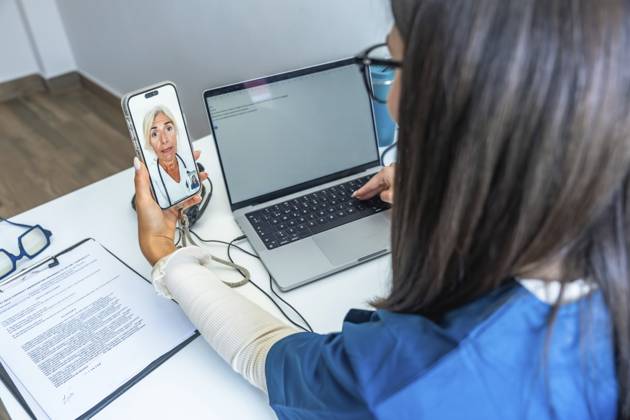 Medizinisches Fachpersonal, das auf einem Smartphone eine Videokonferenz mit einem Arzt abhält und dabei einen Laptop benutzt, um moderne Telegesundheitsdienste und digitale medizinische Kommunikation darzustellen Healthcare professional having a video conference with a doctor on a smartphone while using a laptop, representing modern telehealth services and digital medical communication Copyright: imageBROKER UnaixHuizi ibxuhu15445167.jpg ,model released, Symbolfoto ,property released