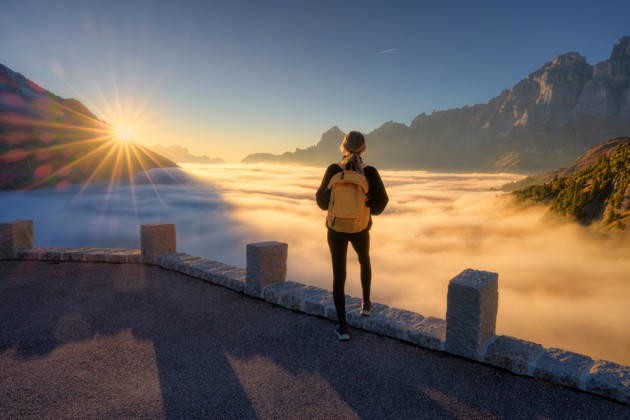 Woman with yellow backpack stands on a viewpoint above a sea of clouds at sunset in Swiss Alps. Girl, sun rays illuminate the alpine mountain peaks and misty valley. Travel in Switzerland. Hiker Model Released DenisBilytskyi_ID32879_1119722_019 Copyright: xx