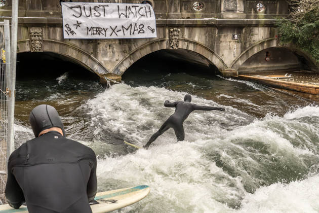 Eisbachwelle ist an Weihnachten zurück, ein Weihnachtswunder, seit Wochen war Stillstand, nun haben sich die Surfer selbst geholfen, provisorisch ein Brett ins Wasser gehängt, die Surfer sind glücklich, München, 26. Dezember 2025 Deutschland, München, 26. Dezember 2025, die Eisbachwelle ist zurück, seit gestern können wieder Surfer auf der Welle surfen, die berühmte Eisbachwelle wurde von den Surfern heimlich selber repariert, zwei Seile sind an der Brücke befestigt, dazwischen hängt ein Brett, das unter Wasser für den nötigen Widerstand sorgt und damit die neue alte Surfwelle aufbaut, Transparent an der Brücke: Just Watch, Merry X-mas , ein Weihnachtswunder, eine große Überraschung zu Weihnachten, seit fast zwei Monaten war