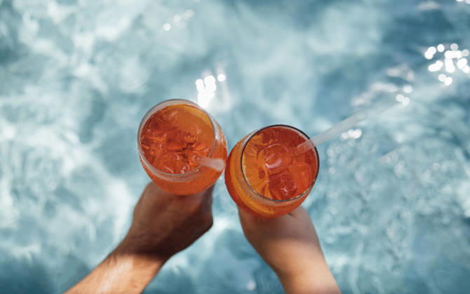 Zwei Hände halten Gläser mit orangefarbenen Cocktails mit Eiswürfeln, die teilweise in einem klaren Swimmingpool eingetaucht sind, Sonnenlicht reflektiert auf der Wasseroberfläche. Two hands grasp glasses of aperol spritz filled with orange cocktails and ice, partially submerged in a clear pool, with sunlight creating reflections on the water (License=RF) Copyright: xZoonar.com DashaxPetrenkox 24646524