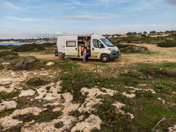 Campers sit in the sliding door of their camper on an unpaved off-road pitch on the Mediterranean coast near Taranto Salento, Puglia, Italy A camper van is parked on a gravel pitch on the shore of the Mediterranean Sea
