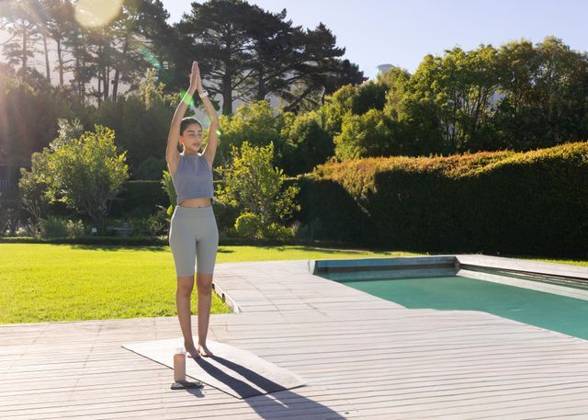 Indian woman practicing yoga in sportswear on wooden deck beside pool with mat, bottle, smartphone, model released, property released, 10.04.2025 06:00:00, 13157950 Copyright: xIMS-260421_4xcreatorid:x166x 13157950