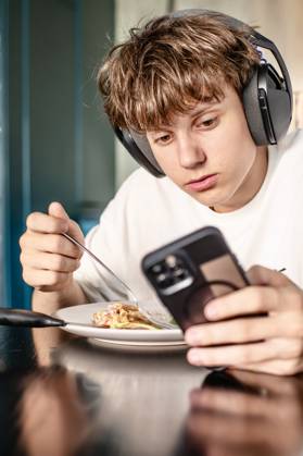 Ein Teenager mit Kopfhörern, der beim Mittagessen zu Hause auf sein Smartphone schaut Focused teenage boy wearing headphones uses his smartphone while having a meal at the kitchen table, distracted by digital content. (License=RF) ,model released, Symbolfoto Copyright: xZoonar.com LiudmylaxLazoryshynax 25142488 ,model released, Symbolfoto ,property released