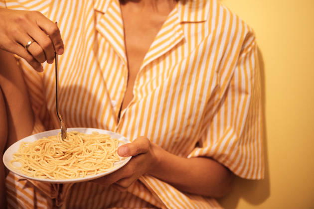 Eine Frau in einem gestreiften Pyjama sitzt an einer gelben Wand und hält einen Teller Spaghetti mit einer Gabel in der Hand; die Szene vermittelt den Eindruck eines ungezwungenen Essens zu Hause Woman in striped sleepwear enjoys a plate of spaghetti while seated against a yellow wall, creating a cozy home dining atmosphere (License=RF) ,model released, Symbolfoto Copyright: xZoonar.com DashaxPetrenkox 25184784 ,model released, Symbolfoto ,property released