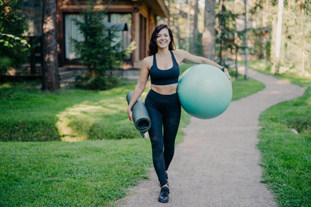 Eine lächelnde Frau trägt einen Gymnastikball und eine Trainingsmatte und geht auf einem Weg in der Nähe eines Holzhauses im Wald spazieren Smiling woman carries fitness ball and exercise mat walking along path near wooden house in forest (License=RF) ,model released, Symbolfoto Copyright: xZoonar.com ViorelxKurnosovx 25185751 ,model released, Symbolfoto ,property released