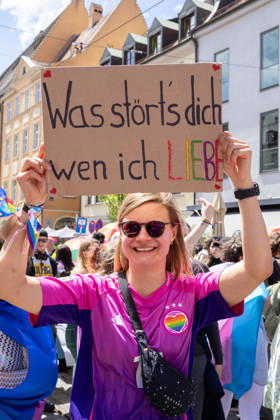 Augsburg, Bavaria, Germany - June 13, 2026: Participants carrying rainbow flags and wearing colorful costumes take part in Christopher Street Day (CSD) in Augsburg. The parade through the city center promotes the rights of LGBTQ+ people as well as social diversity and tolerance. A woman holds a homemade sign in rainbow colors that reads: WHAT™S IT TO YOU WHO I LOVE