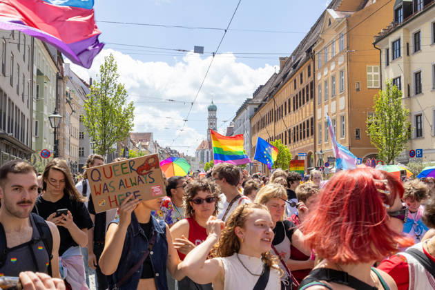 Augsburg, Bavaria, Germany June 13, 2026: Participants carrying rainbow flags and wearing colorful costumes take part in Christopher Street Day (CSD) in Augsburg. The parade through the city center promotes the rights of LGBTQ+ people as well as social diversity and tolerance. A participant holds a sign reading: QUEER WAS ALWAYS HERE