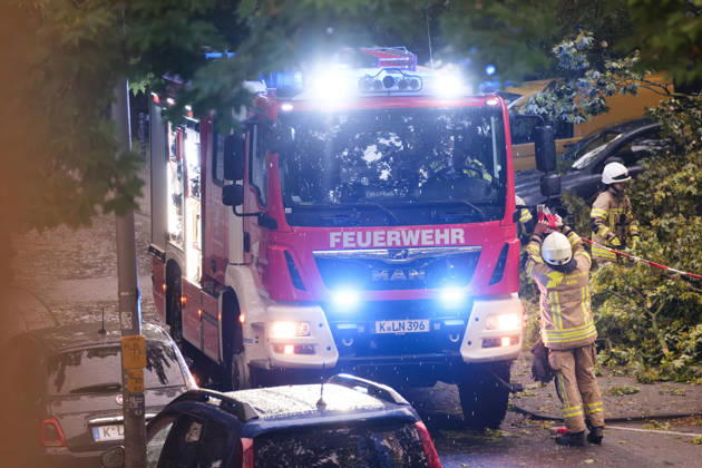 A Cologne fire department truck with flashing blue lights during a continuous emergency operation in Cologne-Nippes; firefighters clearing fallen trees and blocked roads after a severe storm with heavy thunderstorms and gales. Nordrhein-Westfalen Deutschland, Germany DSCF6999