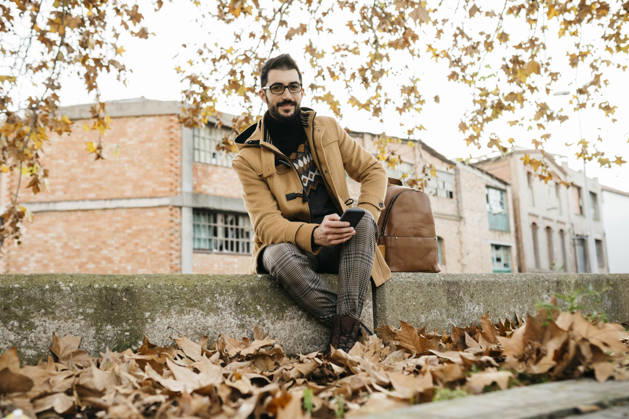 Spain, Igualada, portrait of smiling man with cell phone sitting down in the autumnal town model released, Symbolfoto, JRFF02297