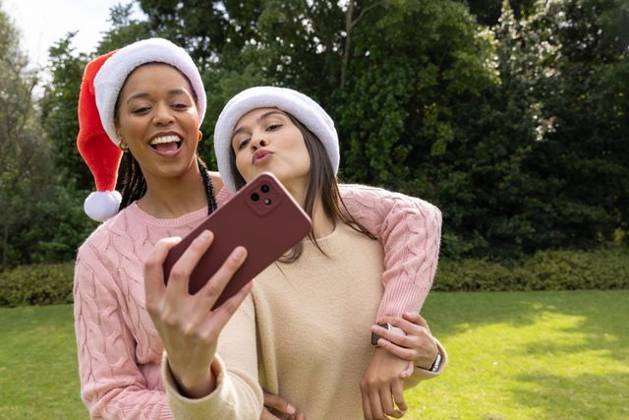 Diverse female friends posing for selfie in park holding smartphone wearing Santa hats and sweaters, model released, property released, 14.08.2025 06:00:00, 7811309 Copyright: xIDS-260430_3xcreatorid:x0x 7811309