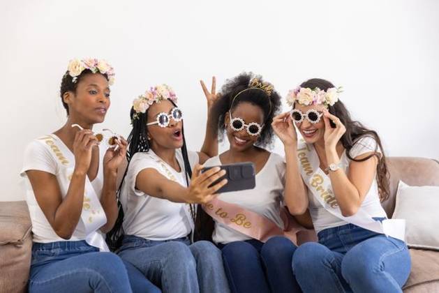 Diverse female friends posing for selfie on sofa wearing sashes sunglasses while holding smartphone, model released, property released, 29.11.2024 06:00:00, 6581265 Copyright: xIDS-250922xcreatorid:x0x 6581265