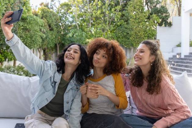 Diverse female friends posing for selfie on white outdoor sofa, holding smartphone and pink mug, model released, property released, 02.09.2025 06:00:00, 8094331 Copyright: xIDS-260501xcreatorid:x0x 8094331