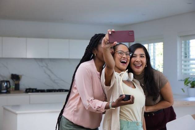 Diverse female friends posing for selfie in modern kitchen, holding red smartphone and headphones, model released, property released, 23.04.2026 06:00:00, 16412756 Copyright: xIDS-260911xcreatorid:x0x 16412756