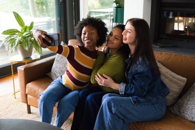 Diverse female friends posing for selfie on tan leather sofa in living room with smartphone, model released, property released, 21.05.2026 06:00:00, 16472399 Copyright: xIDS-261006_2xcreatorid:x0x 16472399