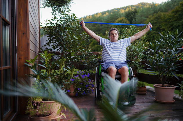 Senior man in wheelchair exercising with resistance band on terrace. Senior man with physical disability exercising independently on terrace. model released