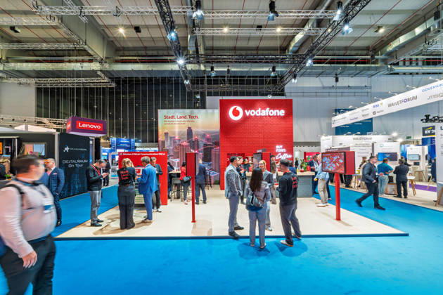Berlin, 13.10.2026: Besucher am roten Messestand von Vodafone unter dem Motto Stadt. Land. Tech. auf der Smart Country Convention; der Telekommunikationskonzern wirbt dort für digitale Infrastruktur für Kommunen und für einsatzkritische Kommunikation (Mission Critical Communication), daneben die Stände von Lenovo und des Walletforums. Die Smart Country Convention (SCCON) ist die Kongressmesse für die Digitalisierung des öffentlichen Sektors und wird vom Digitalverband Bitkom gemeinsam mit der Messe Berlin veranstaltet. Vom 13. bis 15. Oktober 2026 treffen sich auf dem Berliner Messegelände Vertreter aus Bund, Ländern und Kommunen mit Unternehmen der Digitalwirtschaft, um über digitale Verwaltung, Smart City und digitale Souve