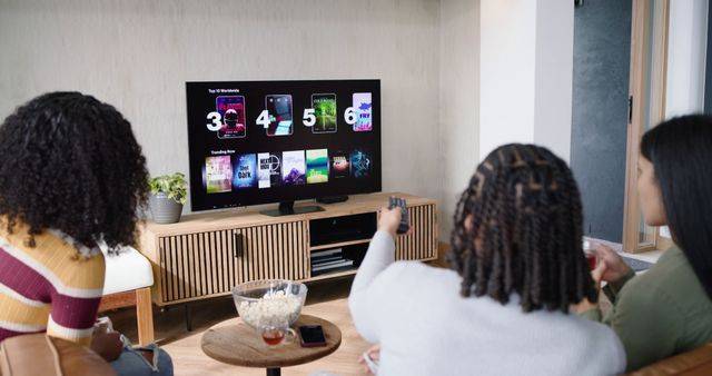 Watching three women in sweaters sitting on sofas pointing remote at TV in living room, popcorn, model released, property released, Cape Town, South Africa, 04.08.2026 06:00:00, 18090323 Copyright: xIPW-261017xcreatorid:x461x 18090323