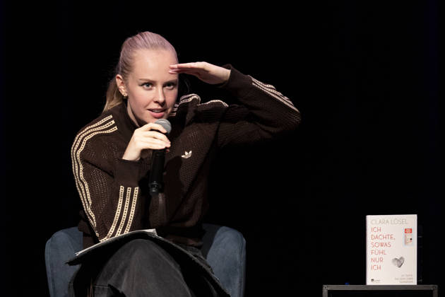 Poetry artist and author Clara LOESEL LÖSEL gazes searchingly into the audience during her reading—a preview show for her new book, *Ich dachte, sowas fühle nur ich*—seated with a microphone, a single frame, a single photo, at the Theater am Tanzbrunnen, Cologne, on October 23, 2026