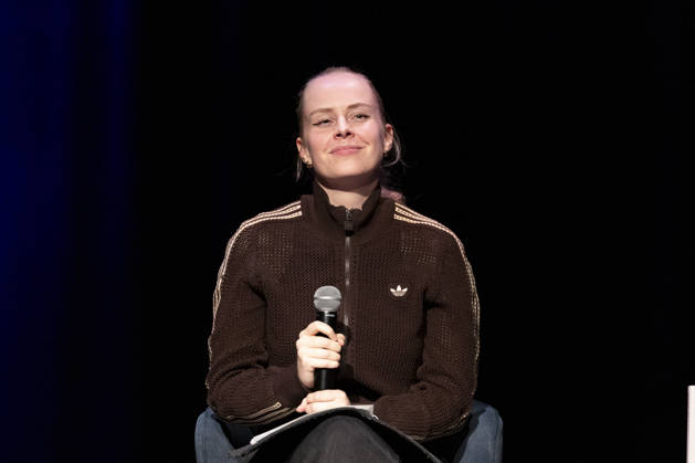 Poetry artist and author Clara LOESEL LÖSEL, smiling, savors the moment during her reading—a preview of her new book, *I Thought I Was the Only One Who Felt This Way*—sitting with a microphone, a single frame, a single photo, at the Theater am Tanzbrunnen, Cologne, on October 23, 2026