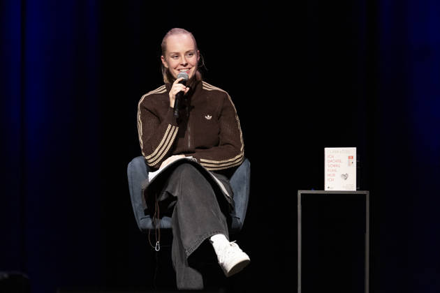 Poetry artist and author Clara LOESEL LÖSEL laughing during her reading, a preview show for her new book *Ich dachte, sowas fühle nur ich* (I Thought I Was the Only One Who Felt That Way), seated with a microphone; single frame, single photo, at the Theater am Tanzbrunnen, Cologne, on October 23, 2026