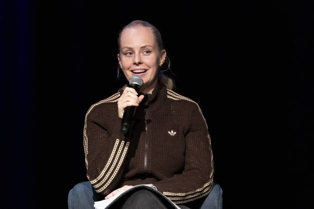 Poetry artist and author Clara LOESEL LÖSEL, smiling and joyful, during her reading—a preview event for her new book, *Ich dachte, sowas fühle nur ich*—seated with a microphone; single frame, single photo, at the Theater am Tanzbrunnen, Cologne, on October 23, 2026