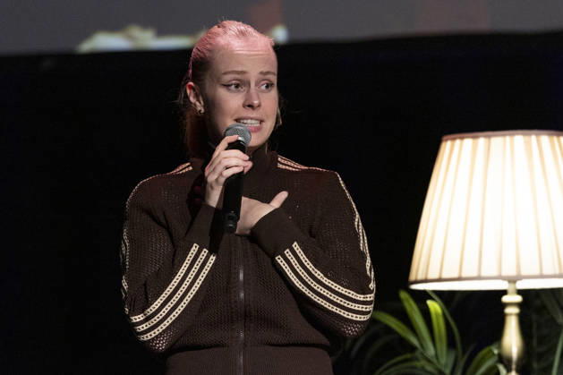 Poetry artist and author Clara LOESEL LÖSEL speaks intently during her reading—a preview of her new book, *Ich dachte, sowas fühle nur ich*—standing with a microphone, in a single frame, a single photograph, at the Theater am Tanzbrunnen, Cologne, on October 23, 2026