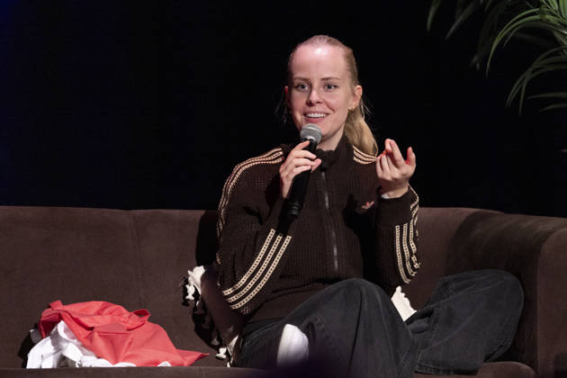 Poetry artist and author Clara LOESEL LÖSEL speaking, gesturing with one hand, during her reading—a preview event for her new book, *Ich dachte, sowas fühle nur ich*—sitting on a sofa with a microphone; single frame, single photograph, at the Theater am Tanzbrunnen, Cologne, on October 23, 2026